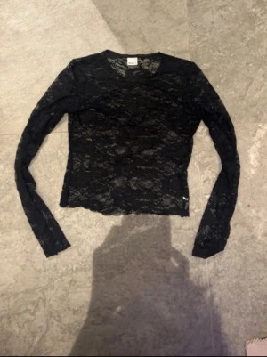 Svart spetsblus från Gina Tricot - Säljer en svart långärmad spetsblus från Gina Tricot. Toppen är genombruten med ett snyggt spetsmönster och har en tight passform. Perfekt att styla med ett linne under eller som lager på lager.