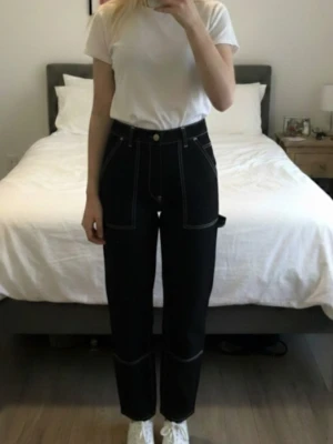 Svarta "snickar" jeans med kontrastsömmar - Svarta jeans med vita kontrastsömmar och stora fickor fram och bak. Byxorna har raka ben och bälteshällor i midjan. Materialet känns slitstarkt och de har en cool, loose passform som ger en avslappnad vibe.