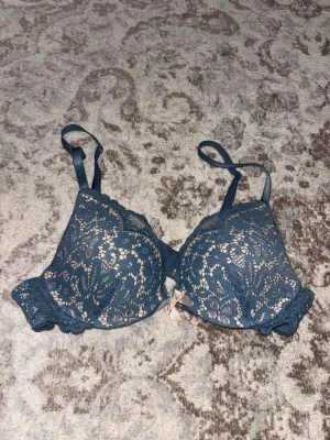 Blå spets-bh med rosett - Snygg blå bh med vadderade kupor och spetsdetaljer över hela framsidan. Bh:n har justerbara axelband och en liten beige rosett framtill. Insidan är beige och materialet är en mix av spets och mjuk textil för extra komfort.