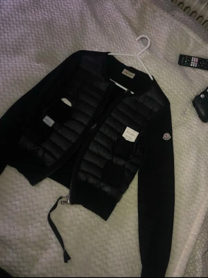 Svart moncler cardigan - Utrustad med NFC-tagg på ärmen samt Certilogo-tagg inuti jackan för äkthetsverifiering.  Skick: Solblekning finns (se bild 2),  Tyget vid dragkedjan har lossnat, så den är i nuläget knuten, Upphängningslappen inuti jackan har spruckna sömmar.  Skriv vid seriöst intresse.