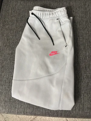 Nike Tech byxor  - Snygga grå mjukisbyxor från Nike med svart dragsko i midjan och rosa Nike-logga på benet. Byxorna har avslappnad passform, sidofickor med dragkedja och är gjorda i mjukt material. Använda ett tag men är i gott skick, 