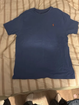 Blå t-shirt från Polo Ralph Lauren - Klassisk blå t-shirt från Polo Ralph Lauren med rund hals och korta ärmar. Liten röd logga broderad på bröstet ger en clean och stilren look. Tillverkad i mjuk bomull som känns skön mot huden. Perfekt till jeans eller shorts. Vet inte riktigt storleken på tröjan, men skulle gissa på S eller XS