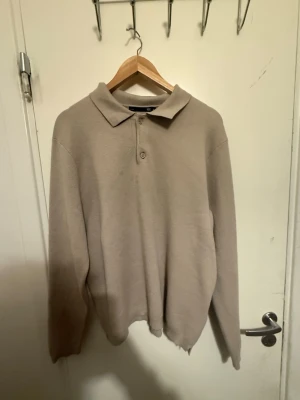 Beige pikétröja med lång ärm - En stilren beige pikétröja med lång ärm och klassisk krage. Tröjan har en knapp framtill och är tillverkad i ett mjukt, tunt material som ger en avslappnad look. Perfekt för dig som gillar minimalistisk stil. Den ser använd och fläckig men de va ljuset 