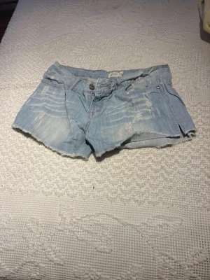 Low waist vintage jeansshorts - Säljer ett par ljusblå jeansshorts med slitna detaljer och rå kant nertill. Klassisk femficksmodell med knapp och dragkedja framtill. Perfekta för sommaren och ger en avslappnad vibe.