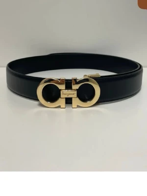 Svart skärp från Salvatore Ferragamo - Stilrent svart skärp från Salvatore Ferragamo med ikoniskt guldspänne i metall. Skärpet är tillverkat i slätt skinn och har en elegant, klassisk design som lyfter vilken outfit som helst.