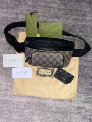 Gucci magväska med monogram - Säljer en snygg magväska från Gucci i beige och svart med klassiskt GG-monogram. Väskan har justerbart svart band, dragkedja upptill och en mindre ficka framtill. Tillbehör som äkthetskort och dustbag medföljer. Perfekt accessoar för dig som gillar lyxiga detaljer.