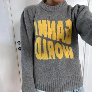 Grå stickad Ganni-tröja med tryck - Säljer en grå stickad tröja från Ganni med stort gult 'GANNI WORLD'-tryck på bröstet. Tröjan har ribbad hals, muddar och nederkant samt långa ärmar med lätt puff vid axlarna. Perfekt för dig som gillar statement-plagg och coola detaljer.