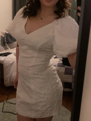 Vit kort klänning med puffärm - Trendig vit kort klänning med puffärmar och v-ringning. Klänningen har en strukturerad yta och figurnära passform som ger en snygg siluett. Perfekt för student eller sommarens alla tillfällen! Den är använd en gång under min studentdag och den var perfekt! Bilden när jag sitter ser den lite kort ut för att den inte var helt helt neddragen, men överlag är den perfekt längd!🤩