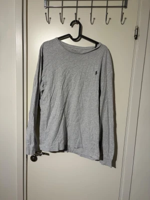 Polo Ralph Lauren tröja   - Ljusgrå långärmad tröja från Polo Ralph Lauren med rund halsringning och den klassiska lilla broderade loggan på bröstet. Tillverkad i mjuk bomull, perfekt för en avslappnad och stilren look.