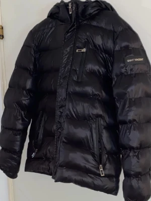 Svart dunjacka från Saint Vincent - Svart dunjacka med quiltad design från Saint Vincent. Jackan har huva, långa ärmar och flera dragkedjeförsedda fickor. Perfekt för kalla dagar och har en modern, lite loose passform. Stilsäker och funktionell för vinter. Den har ett litet hål i fickan vid bröstet men inget som märks så pass mycket. Pris kan diskuteras 