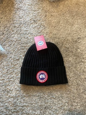Svart ribbad mössa från Canada Goose - Säljer en svart ribbstickad mössa från Canada Goose med klassisk uppvikt kant och det ikoniska runda märket framtill. Perfekt för kalla dagar och enkel att matcha med olika outfits.