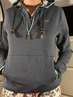 Mörkgrå hoodie med dragkedja - Snygg mörkgrå hoodie från Ragwear med ribbad struktur, turkos dragkedja och detaljer i läderimitation. Modellen har huva med dragsko och känguruficka framtill. Perfekt för en chill och avslappnad stil.