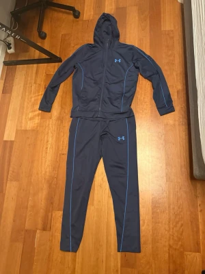 Mörkblå träningsset från Under Armour - Snyggt mörkblått träningsset från Under Armour med matchande hoodie och byxor. Setet har blå kontrastsömmar och dragkedja på tröjan. Perfekt för träning eller uppvärmning. Materialet är troligtvis polyester för bra andningsförmåga.