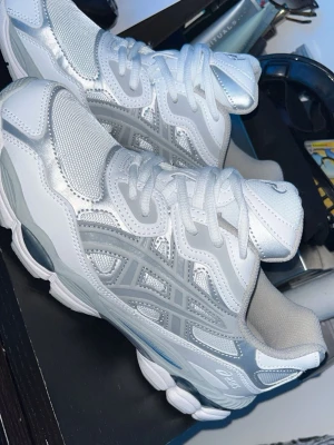 Vita och silver Asics sneakers - Snygga sneakers från Asics i vitt och silver med mesh- och syntetpaneler. Skorna har en chunky sula och klassisk snörning. Perfekta för dig som gillar sportig och trendig stil.