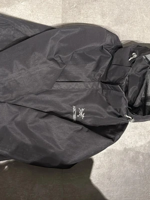 Svart regnjacka från Arc'teryx - Snygg svart regnjacka från Arc'teryx med huva och dragkedja framtill. Jackan har minimalistisk design, justerbar huva och är tillverkad i ett vattentätt material med meshfoder vid halsen. Perfekt för regniga dagar.