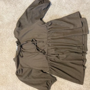 Brun blus med knyt från 157 - Säljer en brun blus från 157 i ribbat material. Blusen har trekvartsärm, puffiga axlar och ett snyggt knytband vid halsen. Den har en avslappnad passform med rynkade detaljer i midjan och vid ärmsluten. Perfekt för en trendig och bekväm look. Liten fläck nedanför bröstet. Se bild! Kanske går att gnugga bort!