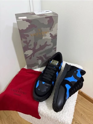 Valentino Garavani blå camo sneakers - Snygga sneakers från Valentino Garavani med blå och svart camo-mönster. Skorna har svarta snören, robust sula och detaljer i mocka och textil. Kommer med originalkartong och röd dustbag. Perfekta för dig som vill sticka ut med exklusiv design.