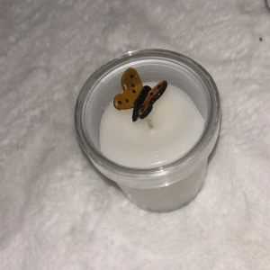 Vit doftljus i glas med fjäril - Säljer ett vitt doftljus i genomskinligt glas med lock. På locket sitter en dekorativ brun och svart fjäril i plast. Perfekt som inredningsdetalj för att skapa mysig stämning i rummet.