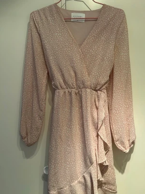 Ljus omlottklänning från Sisters Point - Supersöt ljus kortklänning från Sisters Point med diskret mönster, omlottmodell och volangdetaljer. Klänningen har v-ringning, långa ärmar och ett knytband i midjan som ger en snygg passform. Perfekt för vår och sommar!