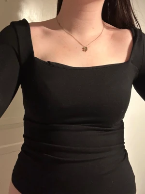 Svart långärmad topp med fyrkantig ringning - Snygg svart topp med lång ärm och fyrkantig ringning både fram och bak. Figurnära passform som framhäver siluetten. Perfekt att styla med jeans eller kjol för en trendig look.