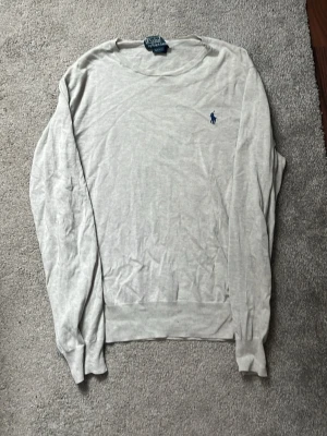 Ljusgrå tröja från Polo Ralph Lauren - Ljusgrå tjocktröja lite tunnare från Polo Ralph Lauren i slim fit-modell. Tröjan har rund halsringning, ribbade muddar och en liten blå broderad logga på bröstet. Perfekt för en stilren och avslappnad look.