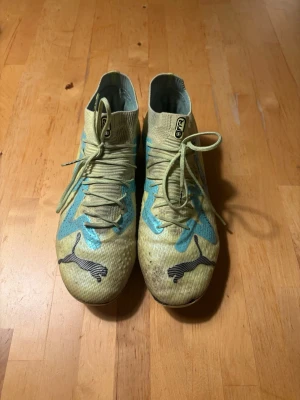 Puma gula fotbollsskor med snörning - Snygga fotbollsskor från Puma i gul färg med ljusblå detaljer och klassisk svart Puma-logga på tån. Skorna har snörning och en högre strumpliknande krage för extra stöd. Perfekta för dig som vill sticka ut på planen.