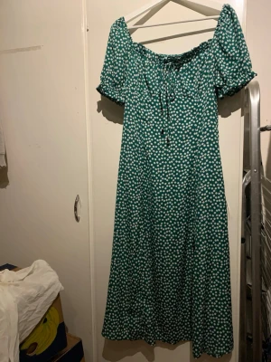 Grön blommig offshoulder klänning SHEIN - Grön långklänning från SHEIN med vita små blommor, offshoulder-modell och korta puffärmar. Klänningen har snörning framtill vid urringningen och är tillverkad i ett lätt syntetmaterial. Perfekt för sommarens alla tillfällen!