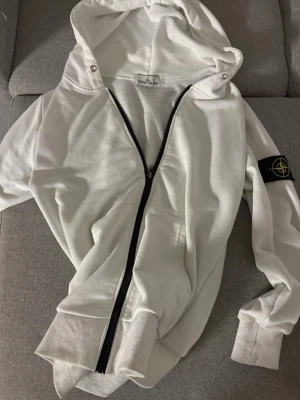Vit hoodie från Stone Island - Säljer en vit hoodie från Stone Island med dragkedja framtill och klassisk svart logopatch på ärmen. Hoodien har huva med snörning och ribbade muddar vid ärmslut och nederkant. Perfekt för dig som gillar streetwear och vill ha något stilrent.