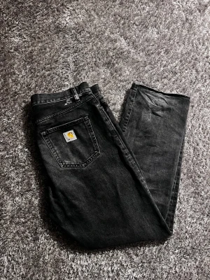 Carhartt jeans  - Säljer ett par svarta jeans från Carhartt med klassisk rak passform. Jeansen har en bakficka med Carhartt-logga och är tillverkade i slitstark denim. Perfekta för dig som gillar streetwear och vill ha ett snyggt basplagg. Storlek 36/32 loose/baggy med skärp passar dem någon som e 185, skick 7/10 en färgfläck som på bild annars bra skick