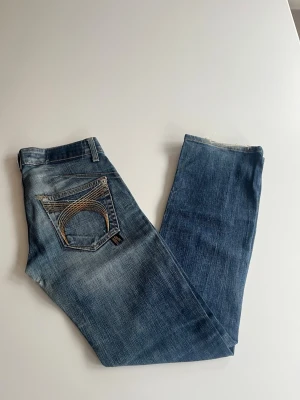 Blå bootcut low waist jeans  - Snygga blå jeans med klassisk gul brodyr på bakfickorna. De har utsvängd passform. Modell: CR 425D 80 DK vintage. Storlek: ca 36 