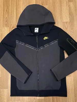 Nike Tech Fleece hoodie grå/svart - Nike Tech Fleece hoodie i grått och svart med dragkedja framtill och huva. Ikonisk gul Nike-logga på bröstet och snygga detaljer på ärmarna. Tillverkad i mjukt och lätt material som passar perfekt till streetwear-stilen.