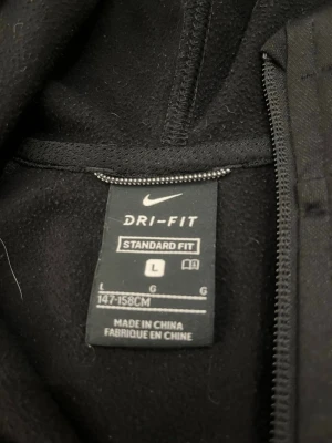 Svart Nike Dri-Fit hoodie med dragkedja - Svart hoodie från Nike med Dri-Fit-material och standard fit. Stor vit Nike-logga på bröstet, dragkedja framtill och två fickor. Huvan är mjuk och skön, perfekt för en sportig och avslappnad stil.