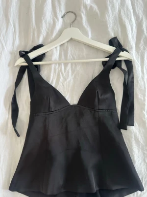 Svart satinlinnetopp med knytband - Stilren svart linnetopp i satin med smickrande v-ringning och breda axelband som knyts på axlarna. Toppen har en lätt utsvängd passform och är perfekt för att skapa en chic look.