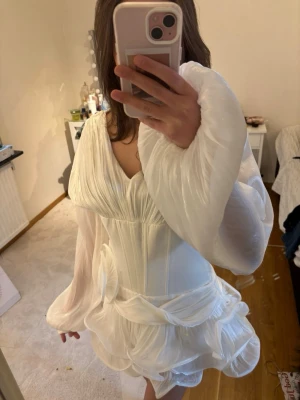 Vit kortklänning med volang och rosett - Supersnygg vit kortklänning med transparenta långärmar, markerad midja och stora volangdetaljer samt en dekorativ rosett framtill. Klänningen har en unik struktur och ger en riktigt statement-look. Perfekt för dig som vill sticka ut! Perfekt till studenten