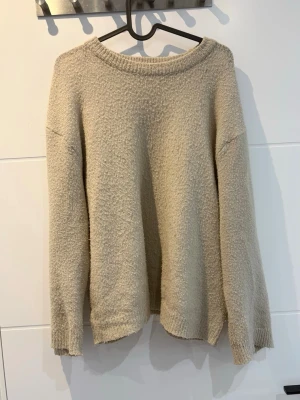 Beige stickad tröja från 157 - Mysig beige stickad tröja från 157 med avslappnad passform och rund halsringning. Tröjan har långa ärmar och ribbade muddar vid hals, ärmslut och nederkant. Perfekt för lager på lager och enkel att matcha med olika outfits.