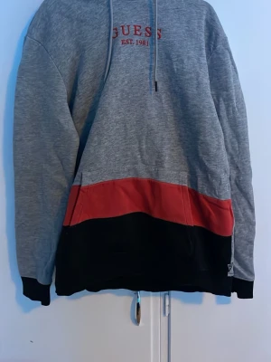 Grå hoodie från Guess med svart och röd detalj - Snygg grå hoodie från Guess med röd och svart färgblock längst ner. Guess-logga och 'Est. 1981' broderat på bröstet. Klassisk huva med dragsko och mjukt material, perfekt för en avslappnad streetwear-look.