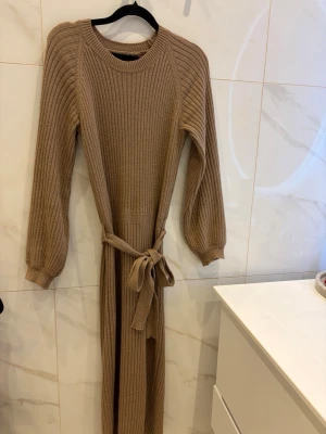 Brun  ribbad långklänning med bälte - Bubbleroom  brun  ribbad långklänning med rund hals, långa ärmar och ett matchande bälte i midjan. Klänningen har en slits på baktill  och är tillverkad i ett mjukt stickat material som ger en skön och följsam passform.