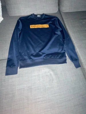 Mörkblå sweatshirt från EA7 Emporio Armani - Snygg mörkblå sweatshirt från EA7 Emporio Armani med orange logga på bröstet och liten logga i nacken. Tröjan har rund hals, långa ärmar och ribbade muddar. Perfekt för en avslappnad och trendig stil.