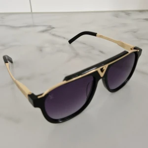 Louis Vuitton svarta solglasögon med gulddetaljer - Snygga solglasögon från Louis Vuitton med svart båge och guldfärgade detaljer på sidorna och överdelen. Glasen är mörka med en lätt lila ton och har LV-loggan ingraverad. Modellen har en kantig och modern form som ger en lyxig vibe. Självklart äkta. Köpta vi Louis Vuitton:↙️ https://en.louisvuitton.com/eng-nl/men/accessories/sunglasses/gold/black/_/N-t194fv1y-axbx9vr3_pb5r44?page=3.                                           Nypris: 6250:-. Kvitto skickas när köpet är färdigt. Skick 8 av 10.