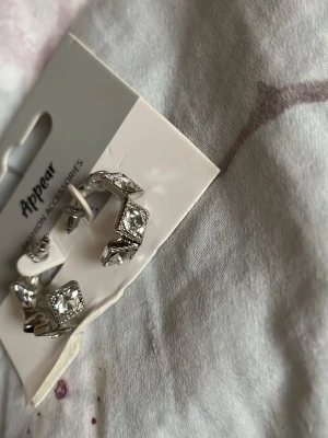 Silvriga örhängen med stora stenar - Säljer ett par silvriga örhängen från Appear. De är halvcirkelformade och dekorerade med stora, fyrkantiga glittrande stenar som ger en lyxig känsla. Perfekta för dig som vill ha något som sticker ut och glänser lite extra.