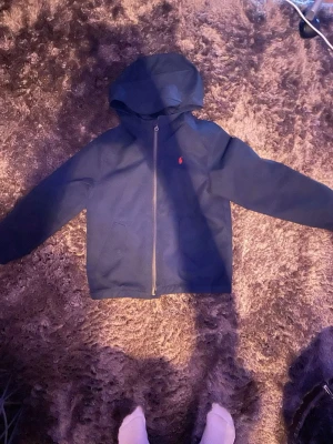 Marinblå vindjacka från Polo Ralph Lauren - Snygg marinblå vindjacka från Polo Ralph Lauren med huva och dragkedja framtill. Jackan har två fickor med lock och den klassiska röda logotypen broderad på bröstet. Perfekt för blåsiga dagar och enkel att matcha med olika outfits. Storlek 8a men passar jag som är 150cm