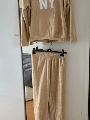 Beige loungewear mjukisdress - Supermysigt beige loungewear-set i fluffigt material. Tröjan har långa ärmar och vitt NY-tryck framtill, byxorna har resår i midjan för extra komfort. Perfekt för chill dagar hemma eller när du vill vara extra bekväm.