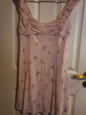 Ljusrosa blommig kortklänning - Söt ljusrosa kortklänning med små blommor i olika färger. Klänningen har breda axelband med rynk och en lätt rynkad byst. Perfekt för varma dagar och har en luftig, feminin känsla.