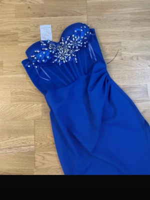 Blå axelbandslös balklänning med pärlor - Axelbandslös blå långklänning från Jolly Glamour med glittriga paljetter och pärlor över bysten. Klänningen har en elegant drapering och transparenta meshdetaljer upptill. Perfekt för bal eller festliga tillfällen.