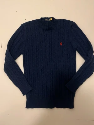 Mörkblå kabelstickad tröja Polo - Mörkblå kabelstickad tröja från Polo Ralph Lauren med rund halsringning och röd broderad logga på bröstet. Tröjan har långa ärmar och ribbade muddar. Perfekt för lager-på-lager och klassisk stil.