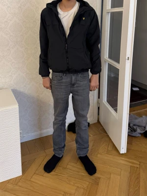 Svart vindjacka från Lyle & Scott - Snygg svart vindjacka från Lyle & Scott med dragkedja framtill och huva. Jackan har en gul logga på bröstet och elastiska muddar vid ärmar och midja. Perfekt för blåsiga dagar och enkel att matcha med olika outfits.