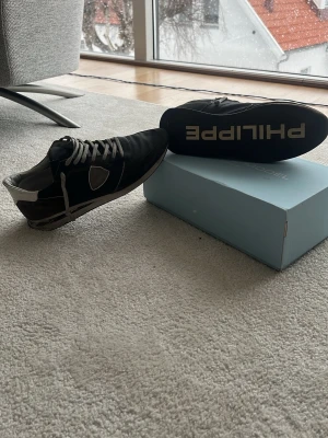 Svarta Philippe Model sneakers - Snygga svarta sneakers från Philippe Model med vita detaljer och gråa skosnören. Skorna har en låg silhuett och är tillverkade i mocka och läder. Klassisk logga på sidan och Philippe-tryck under sulan. Perfekta för en trendig streetwear-look.