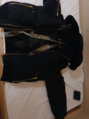 Svart dunjacka från Moose Knuckles - Snygg svart dunjacka från Moose Knuckles med guldfärgade dragkedjor och detaljer. Jackan har flera fickor med dragkedja och en stor, fluffig huva med svart fuskpäls. Perfekt för kalla vinterdagar och riktigt varm.