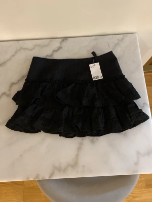 Svart volangkjol med spetsdetaljer - Supersöt svart kortkjol med flera lager volanger och fina spetsdetaljer. Kjolen har en bred midja och är perfekt för dig som gillar en feminin och trendig stil. Passar till både sneakers och boots.