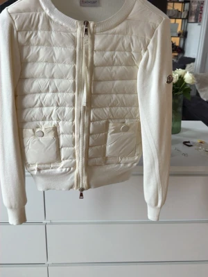 Vit quiltad cardigan från Moncler  - Snygg vit cardiganjacka från Moncler med quiltad front och två fickor med tryckknappar. Jack speeds med dragkedja och har ribbade muddar. Ärmar och rygg i mjukt stickat material, medan framsidan är i glansigt tyg. Moncler-logga på ärmen.